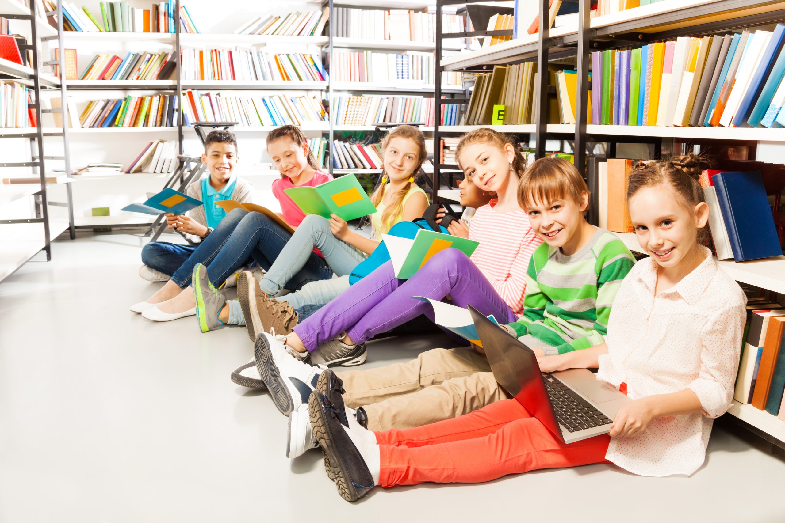 Six smiling children sitting in a row on floor in the library near bookshelf and looking straight